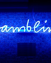 A bright blue neon sign spells out the word 'gambling' in cursive script. The background features a textured wall covered in domino tiles, also illuminated by the blue light, creating a vivid and visually striking scene.