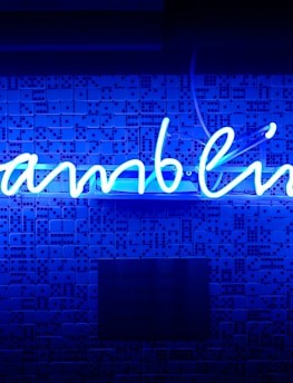 A bright blue neon sign spells out the word 'gambling' in cursive script. The background features a textured wall covered in domino tiles, also illuminated by the blue light, creating a vivid and visually striking scene.