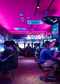 A vibrant arcade with rows of gaming machines adorned with colorful screens. Several people are immersed in playing games, seated on stools in a dimly lit room accented by an intense pink ceiling light. The atmosphere is lively and engaging.