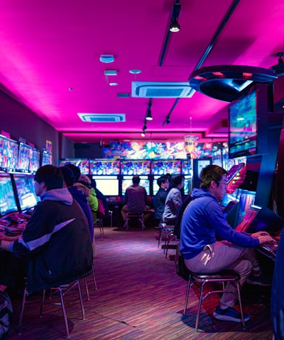 A vibrant arcade with rows of gaming machines adorned with colorful screens. Several people are immersed in playing games, seated on stools in a dimly lit room accented by an intense pink ceiling light. The atmosphere is lively and engaging.