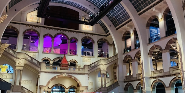 The interior of a large, ornately designed building with arched ceilings and intricate architectural details. The space features gaming machines lined up along the floor, under a high ceiling with a grid pattern. Lighting in purple and white tones enhances the ambiance.