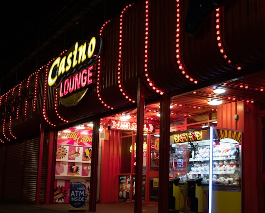 A brightly lit casino lounge entrance adorned with neon lights. The exterior features colorful signage including the words 'Casino' and 'Lounge' in vivid colors, with additional lights framing the building. Inside, a glimpse of a claw machine filled with plush toys and arcade games is visible. The area exudes an inviting yet bustling atmosphere typical of entertainment venues.
