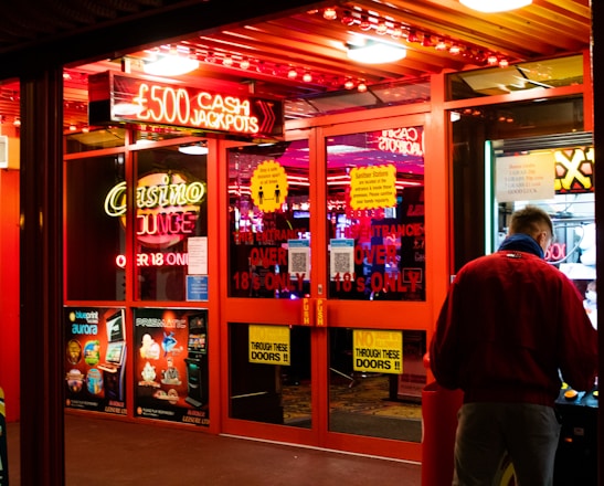 A brightly lit entrance to a casino with neon signs, including one advertising a £500 jackpot. The doors have clear signage indicating entry is restricted to those over 18. A person in a red jacket stands to the right, possibly interacting with a machine or display.